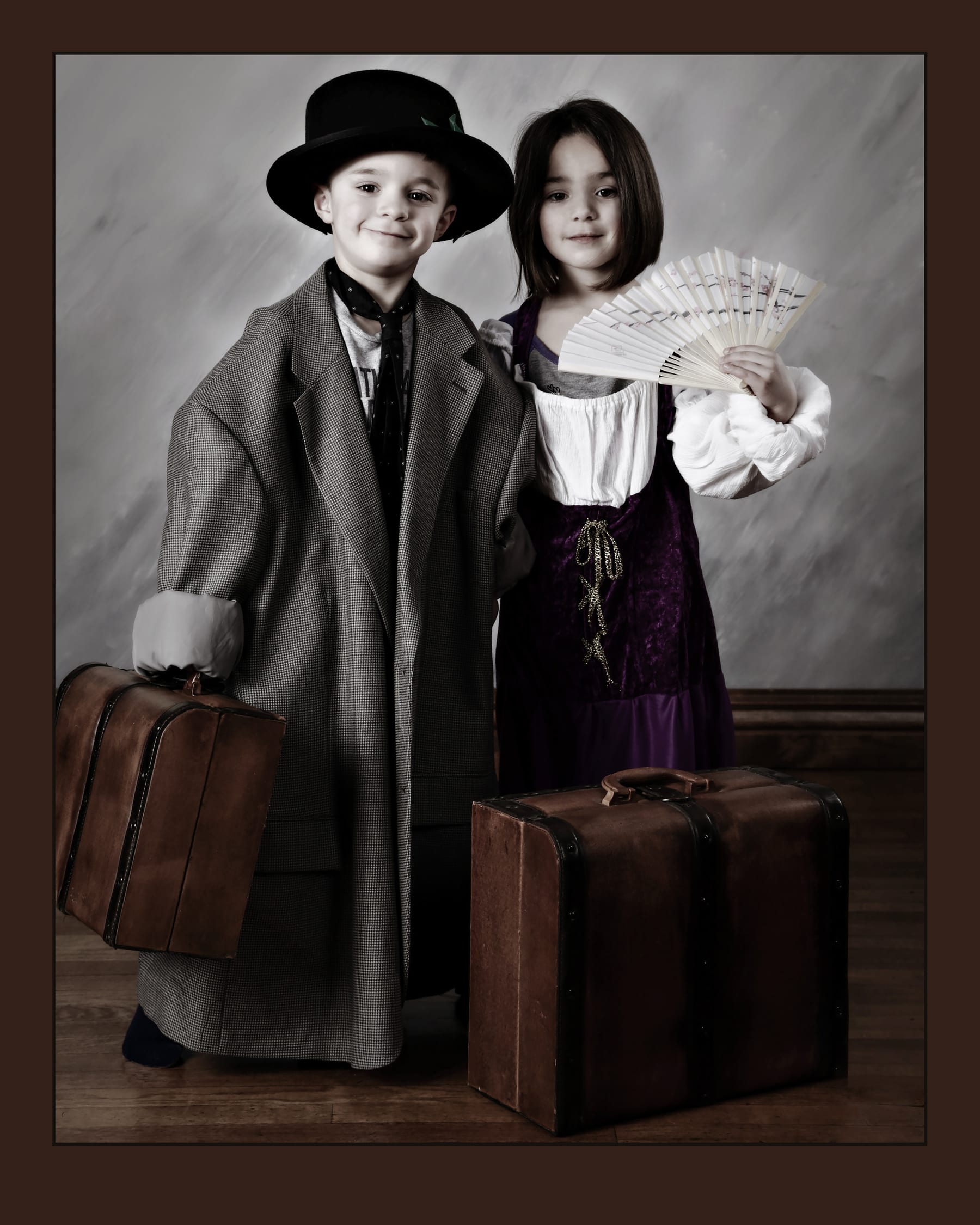 Two children in vintage coats and hats standing beside old leather suitcases in the studio