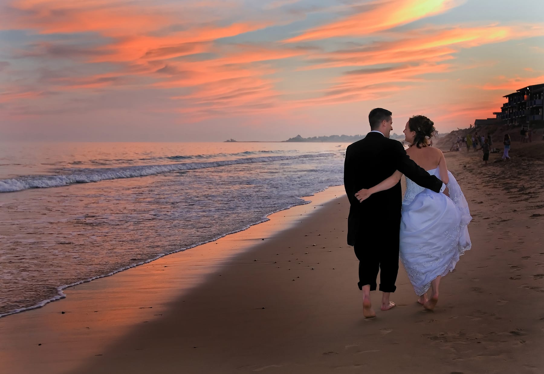 Bride and groom walking barefoot along the shoreline at sunset